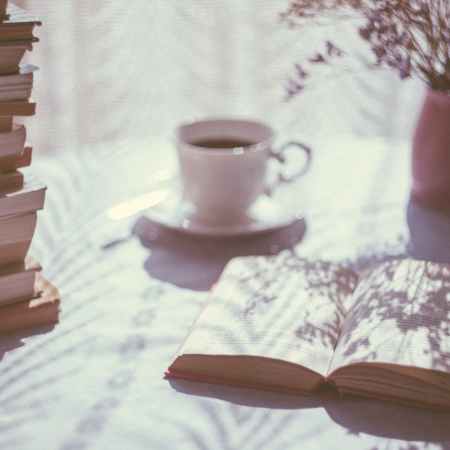 Photo of a table with an open book, a cup of coffee, a vase of flowers and a large stack of books. The photo has a vintage-style filter.