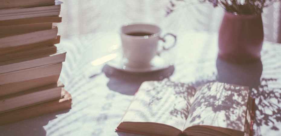 Photo of a table with an open book, a cup of coffee, a vase of flowers and a large stack of books. The photo has a vintage-style filter.