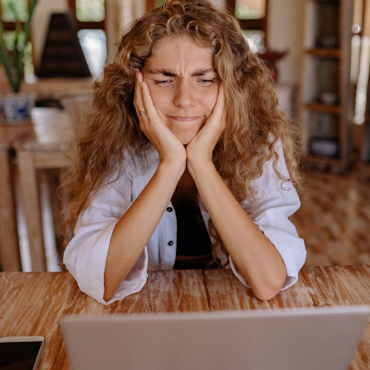 A woman frowning at a laptop screen, with her elbows on the table and her chin resting on her hands, cupping her face.