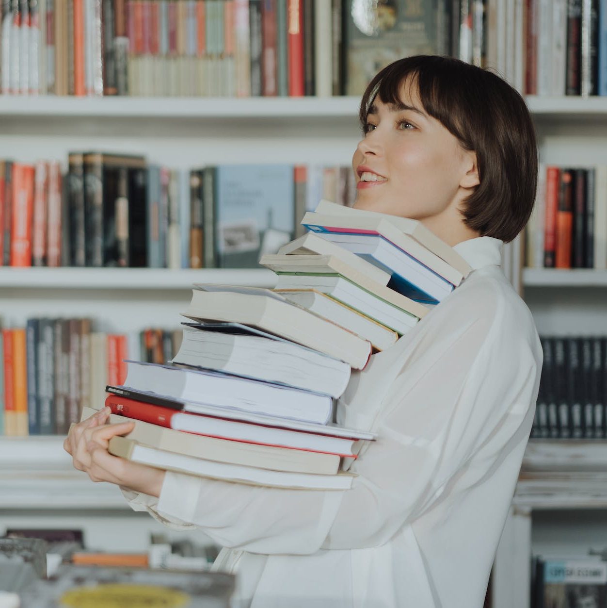 a woman holding a large stack of books that she is holding between her hands and her chin.