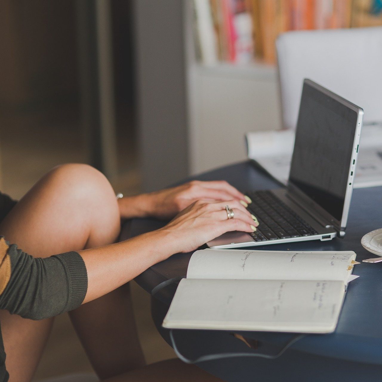 A woman sitting at a table, typing on a laptop with an open notebook next to her. 5 Ways To Find Inspiration For Your Mental Health Blog (wallflowerhaze.com)