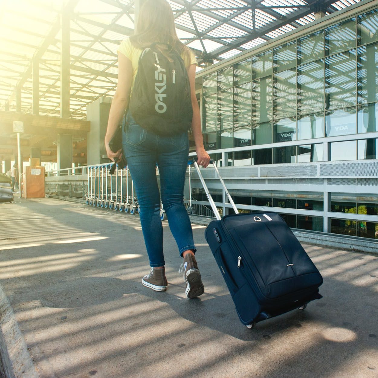 A woman walking away from the camera outside an airport. She is pulling a suitcase behind her and is wearing a backpack. (wallflowerhaze.com)