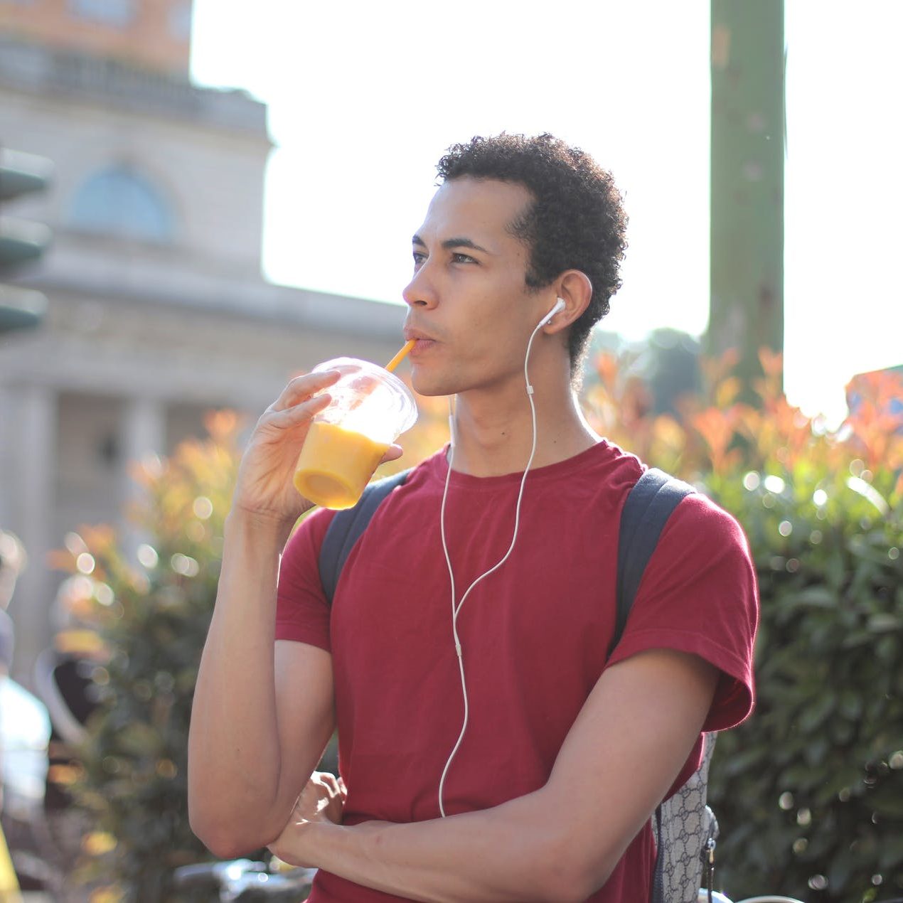 A man standing in the street, drinking from a cup with a straw, and wearing earphones. (wallflowerhaze.com)