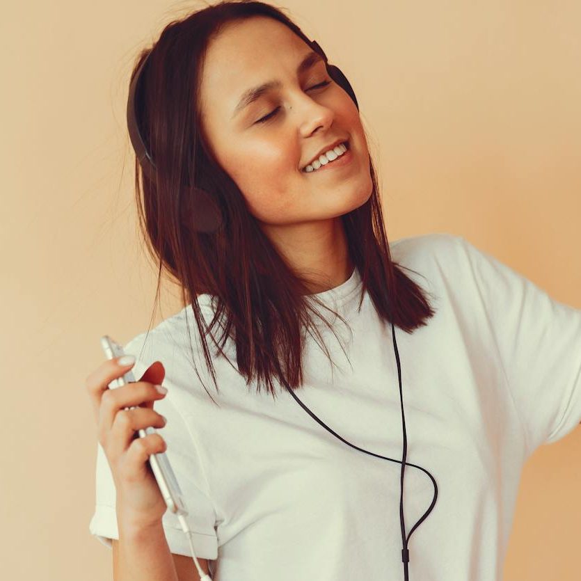 A woman wearing headphones and holding a phone, smiling and dancing whilst listening to music. (wallflowerhaze.com)